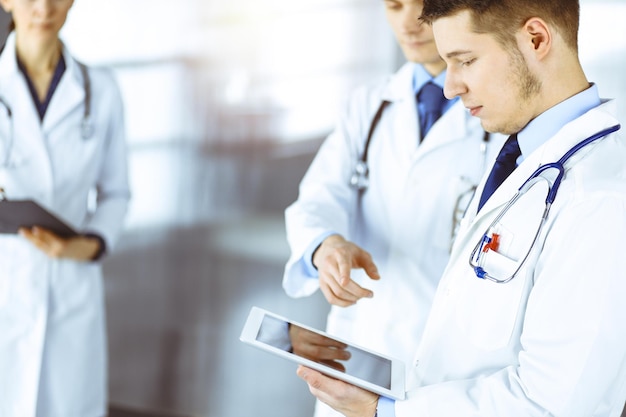 Un grupo de médicos está revisando nombres médicos en una tableta de computadora, con una enfermera usando un portapapeles en el fondo, de pie juntos en una soleada oficina de hospital. Médicos listos para examinar y ayudar