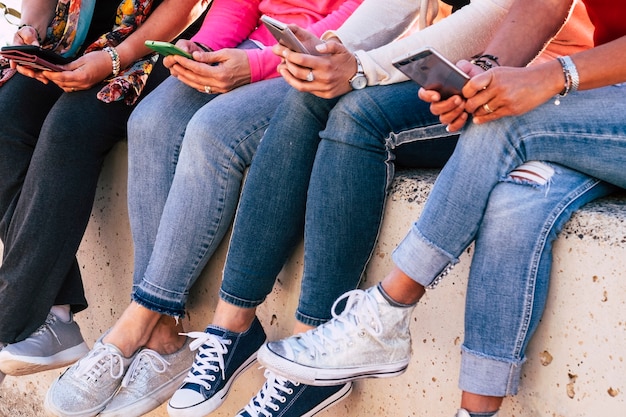 Grupo de mujer casual moderna irreconocible sentada en la pared usando dispositivos telefónicos modernos para redes sociales y medios de Internet - amistad en la forma milenaria moderna