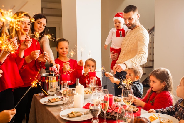 Grupo de mujeres adultas sonrientes felices y hombres parados cerca de la mesa navideña, niñas y niños alegres sentados cerca de los padres con entusiasmo. Destellos, comida sabrosa, champán, decoración navideña.