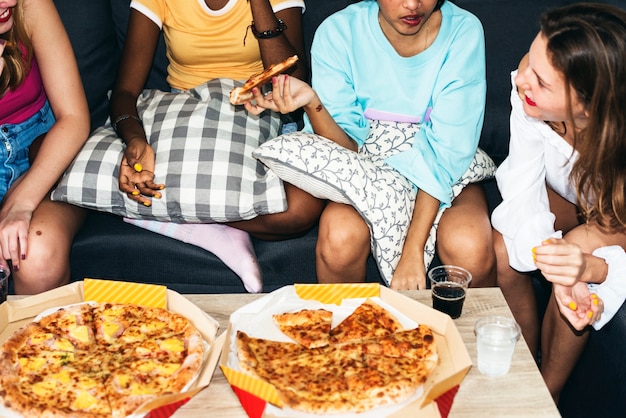 Un grupo de mujeres diversas sentadas en el sofá y comiendo pizza juntos.