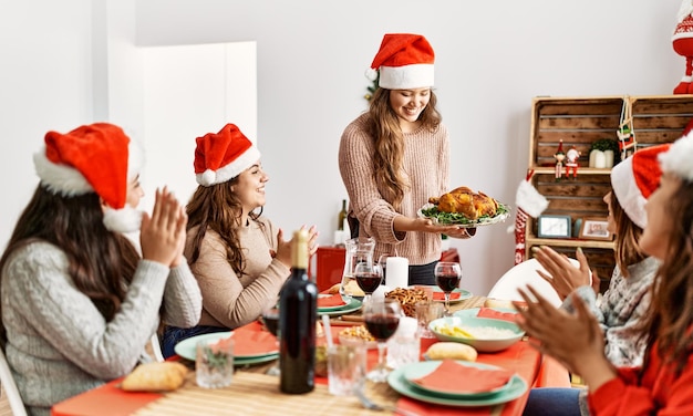 Grupo de mujeres hispanas aplaudiendo y sentadas en la mesa Mujer de pie y sosteniendo pavo asado celebrando la Navidad en casa