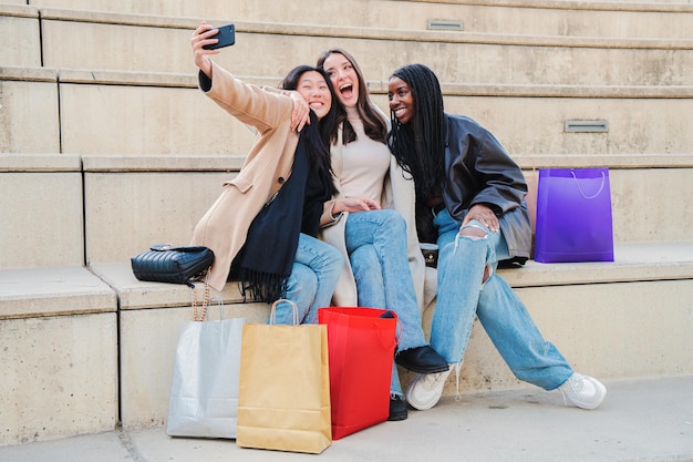 Grupo de mujeres jóvenes multirraciales sonriendo haciendo un retrato selfie con teléfono celular después de comprar Tres chicas felices multiétnicas divirtiéndose tomando fotos sentadas al aire libre Concepto de estilo de vida