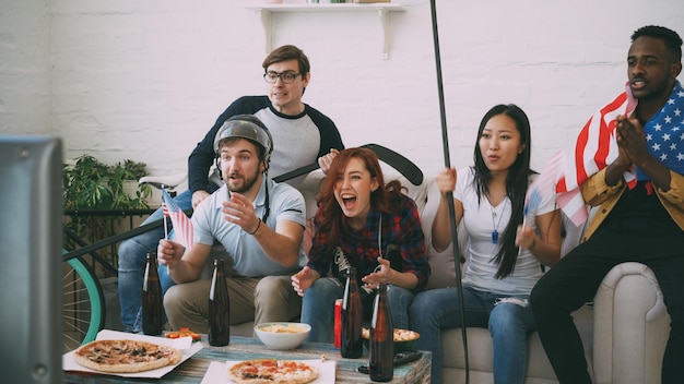 Grupo multiétnico de amigos aficionados al deporte con banderas nacionales de Estados Unidos viendo los juegos olímpicos de invierno