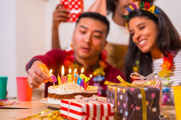 Grupo multiétnico de amigos en una fiesta de cumpleaños en el sofá de casa con un pastel y regalos encendiendo las velas