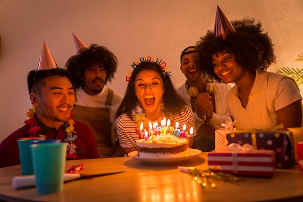 Grupo multiétnico de amigos en una fiesta de cumpleaños en el sofá de casa con un pastel y regalos soplando las velas con las luces apagadas