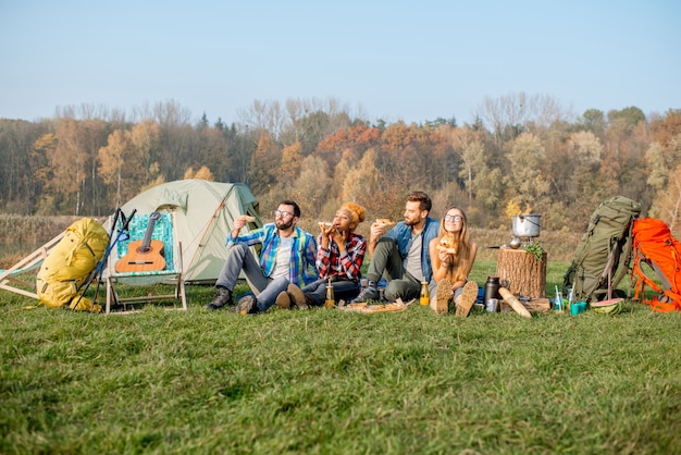 Grupo multiétnico de amigos haciendo un picnic, comiendo pizza, sentados en una fila en el campamento con carpa y equipo de senderismo cerca del bosque