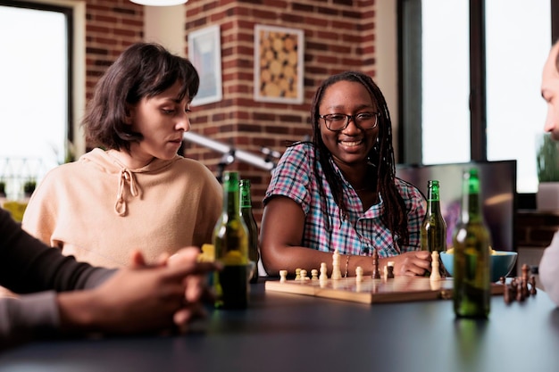 Grupo multiétnico de amigos sentados en casa juntos mientras juegan al ajedrez. Felices personas inteligentes en la sala de estar disfrutando de juegos de mesa estratégicos mientras consumen bocadillos y bebidas.