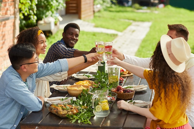 Grupo multiétnico de amigos tintineo de copas de cóctel mientras disfruta de una cena al aire libre en verano