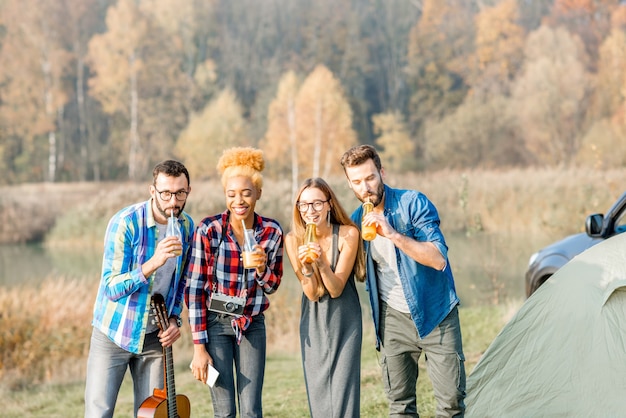 Grupo multiétnico de amigos vestidos casualmente divirtiéndose con bebidas durante la recreación al aire libre en el campamento cerca del bosque