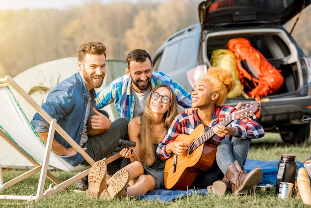 Grupo multiétnico de amigos vestidos casualmente divirtiéndose tocando la guitarra durante la recreación al aire libre con carpa cerca del lago