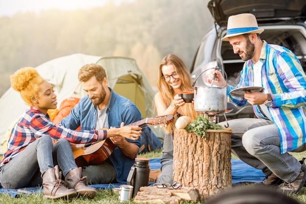 Grupo multiétnico de amigos vestidos casualmente haciendo un picnic, cocinando sopa con caldero, tocando la guitarra durante la recreación al aire libre cerca del lago