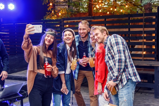 Grupo multiétnico. Chico guapo con barba está haciendo selfie con sus amigos en la fiesta con la cámara de su teléfono. Todos están sonriendo y disfrutando de su compañía, divirtiéndose