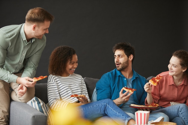 Grupo multiétnico de amigos comendo pizza e lanches enquanto aproveita a festa em casa sentado no grande sofá