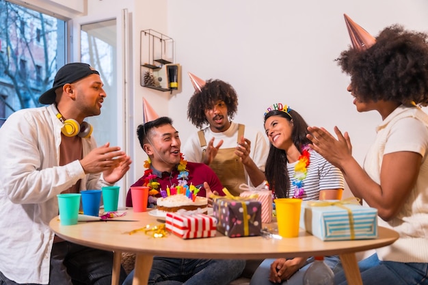 Grupo multiétnico de amigos em uma festa de aniversário no sofá em casa com um bolo e presentes cantando parabéns para ele