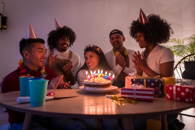 Grupo multiétnico de amigos em uma festa de aniversário no sofá em casa com um bolo e presentes com as luzes apagadas cantando parabéns