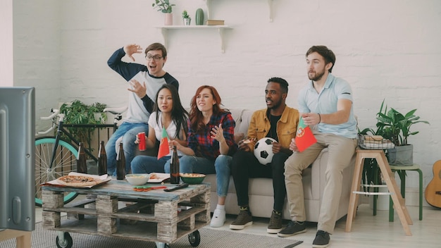 Grupo multiétnico de amigos fãs de esportes com bandeiras portuguesas assistindo campeonato de futebol na TV juntos em casa e animando time favorito