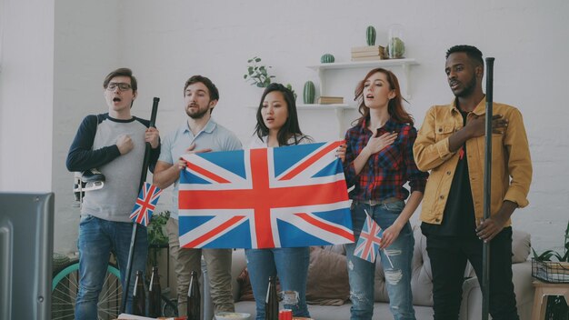 Grupo multiétnico de amigos ouvindo e cantando o hino nacional britânico antes de assistir ao campeonato esportivo na tv juntos em casa