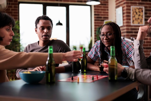 Grupo multiétnico de amigos sentados à mesa enquanto jogavam jogos de tabuleiro com estatuetas. Pessoas diversas felizes desfrutando de jogos da sociedade juntos enquanto comem lanches e bebidas.