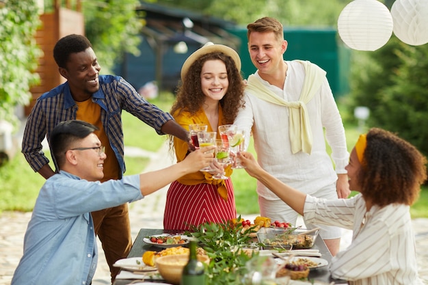 Grupo multiétnico de amigos tilintando copos enquanto desfrutam de um jantar ao ar livre na festa de verão