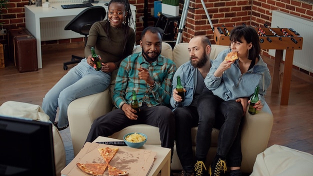 Grupo multiétnico de amigos tilintando garrafas de cerveja na festa em casa, fazendo torradas e assistindo filme na tv. Pessoas felizes curtindo filme no canal de televisão com bebidas alcoólicas.