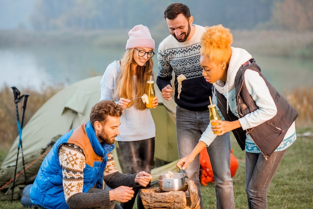 Grupo multiétnico de amigos vestindo suéteres jantando fondue durante a recreação ao ar livre no acampamento à noite