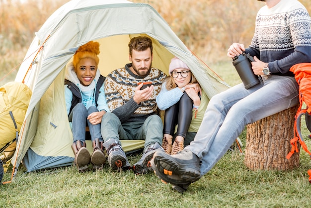 Grupo multiétnico de amigos vestindo suéteres se aquecendo juntos, sentados na tenda durante a recreação ao ar livre perto do lago