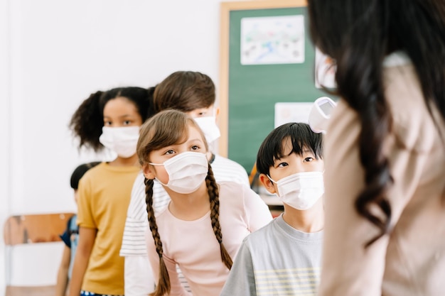 Grupo multiétnico de crianças com máscara facial ficou na fila, deixe o professor verificar a temperatura do termômetro de varredura em busca de febre contra o vírus espalhado na sala de aula após covid19 quarentena e bloqueio