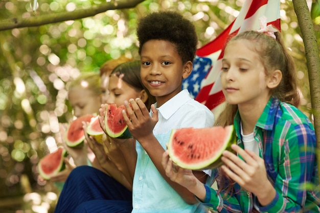 Grupo multiétnico de crianças comendo melancia sentadas em fileira enquanto brincam ao ar livre no verão