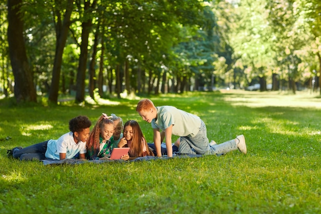 grupo multiétnico de crianças usando tablet digital enquanto deitavam na grama verde em um parque ao ar livre iluminado pela luz solar