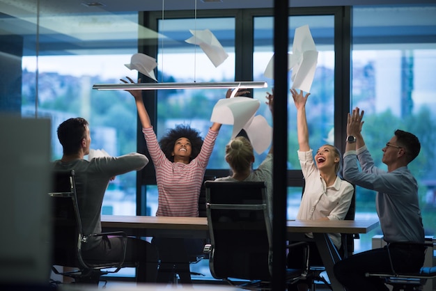 Grupo multiétnico de jovens empresários jogando documentos e parecendo felizes enquanto comemoram o sucesso em seus locais de trabalho no escritório noturno