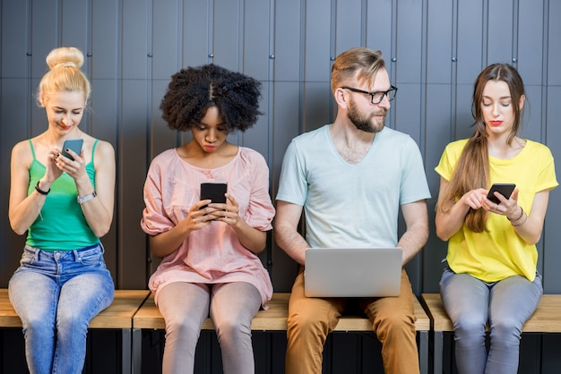 Foto grupo multiétnico de jovens vestidos com camisetas coloridas conversando com gadgets sentados em uma fileira dentro de casa no fundo da parede