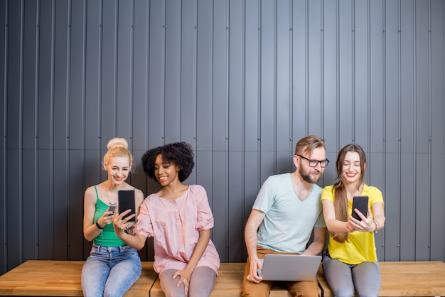 Grupo multiétnico de jovens vestidos com camisetas coloridas conversando com gadgets sentados em uma fileira dentro de casa no fundo da parede