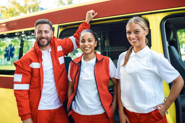 Foto grupo multiétnico de paramédicos de pé ao lado de uma ambulância com as portas abertas seu colega de trabalho carregando um saco de trauma médico eles estão sorrindo para a câmera