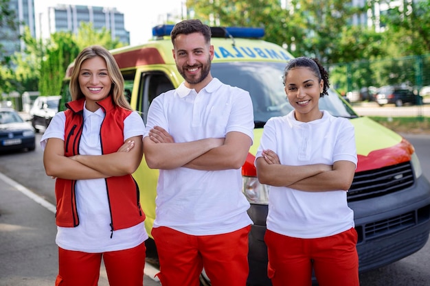 Foto grupo multiétnico de paramédicos de pé ao lado de uma ambulância com as portas abertas seu colega de trabalho carregando um saco de trauma médico eles estão sorrindo para a câmera