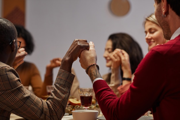 Grupo multiétnico de pessoas de mãos dadas em oração no jantar de ação de graças com amigos e familiares, foco no primeiro plano,