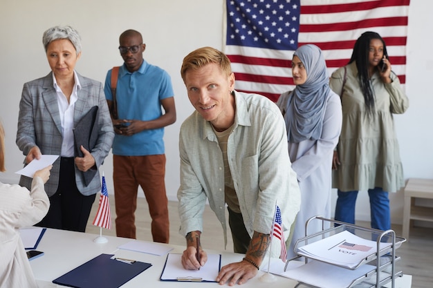 Grupo multiétnico de pessoas que se registram em assembleias de voto decoradas com bandeiras americanas no dia da eleição, foco em homem sorridente assinando boletins de voto e espaço de cópia