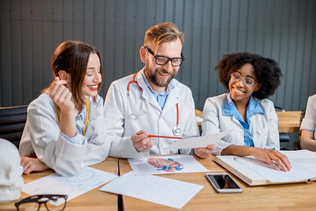 Grupo multiétnico de estudiantes de medicina en uniforme que tienen una discusión sentados juntos en el escritorio con diferentes cosas médicas en el aula