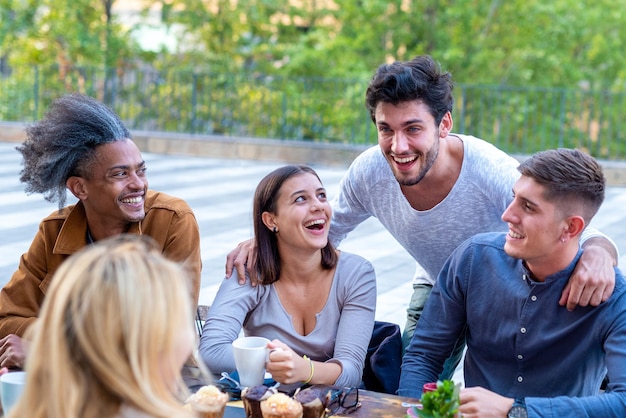 Foto grupo multiétnico de estudiantes universitarios desayunando en la cafetería jóvenes amigos sonriendo y riendo mientras beben café y comen muffins