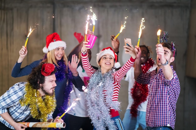 grupo multiétnico de jóvenes empresarios felices e informales tomándose una foto selfie mientras bailan con bengalas y se divierten durante la fiesta de año nuevo frente a un muro de hormigón en una nueva oficina de inicio