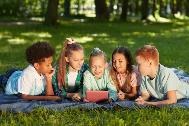 Grupo multiétnico de niños con tableta digital mientras está acostado sobre la hierba verde en el parque al aire libre iluminado por la luz del sol