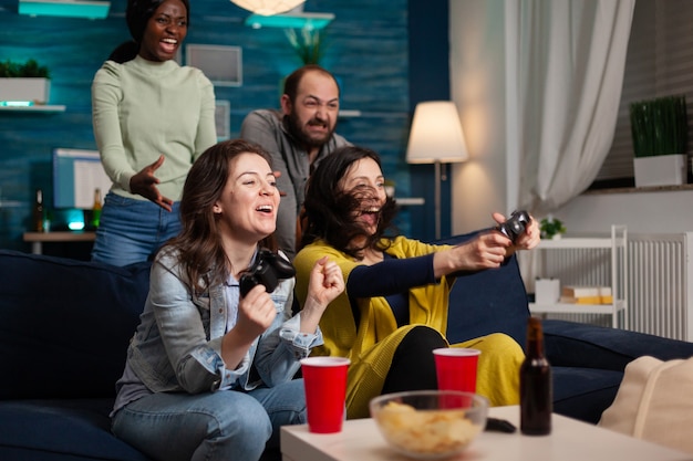 Grupo multiétnico de personas que socializan ganando en videojuegos divirtiéndose. Grupo de amigos de raza mixta jugando juegos mientras está sentado en el sofá en la sala de estar a altas horas de la noche.