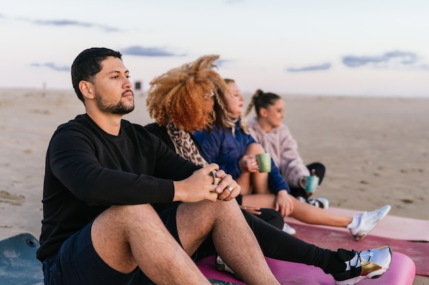 Grupo multiétnico de personas sentadas en esteras en la playa bebiendo té durante la puesta de sol