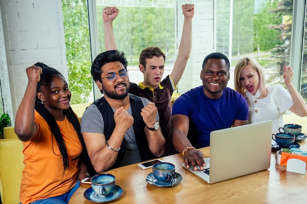 Grupo multinacional de amigos felices disfrutan y beben té mirando el monitor de la computadora portátil y exultantes.