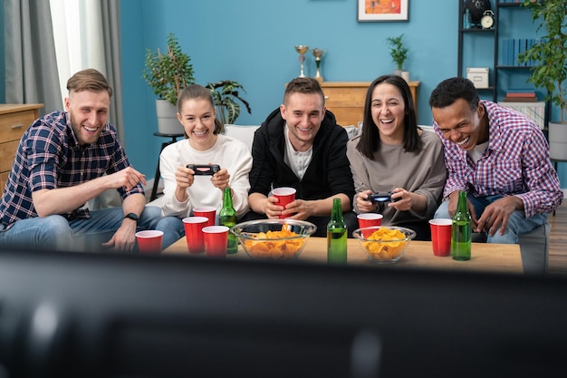 Un grupo multinacional de estudiantes universitarios se sientan en el sofá y juegan videojuegos en la televisión en casa sm