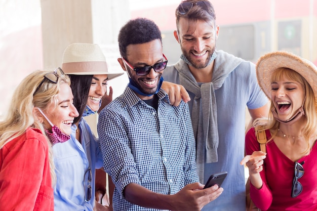 Grupo multirracial de amigos con máscara facial con smartphone en la estación de tren