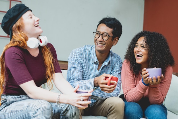 Grupo multirracial de amigos tomando un café juntos, dos mujeres y un hombre riéndose y hablando en la cafetería, gente real divirtiéndose durante el desayuno