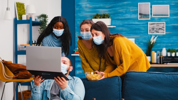 Grupo multirracial de amigos navegando usando laptop, mantendo o distanciamento social e usando máscara facial para evitar disseminação e infecção na sala de estar do apartamento