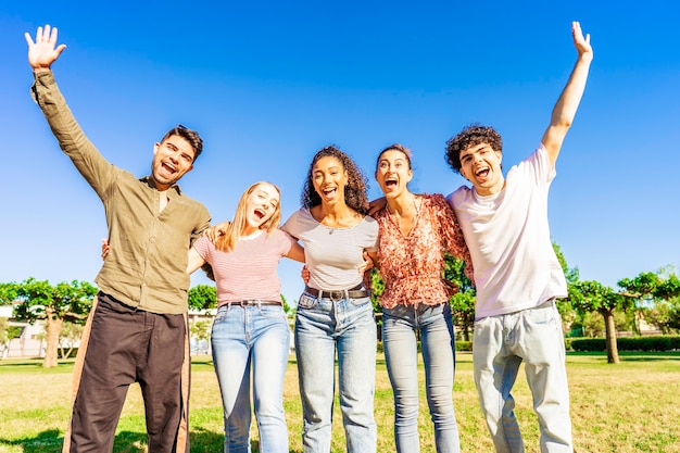 Grupo multirracial de jovens amigos posando com os braços abertos erguidos, olhando para a câmera, abraçando uns aos outros na natureza do parque da cidade. Pessoas felizes e diversificadas se divertindo juntas, curtindo a vida e o sucesso