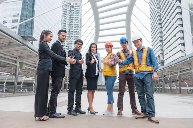 Grupo de negocios multiétnicos con ingeniero de pie y mostrando los pulgares para arriba felicitar el éxito del acuerdo corporativo de cooperación en la ciudad