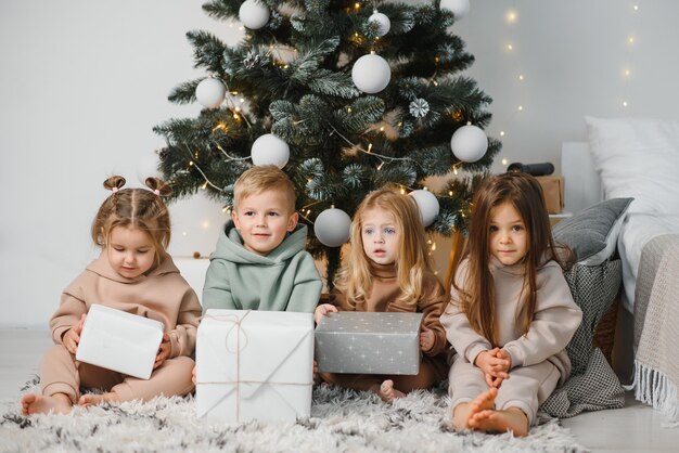 Un grupo de niños bajo un árbol de Navidad en casa. Los niños y niñas con disfraces celebran las vacaciones de invierno. Los niños abren los regalos junto a la chimenea.